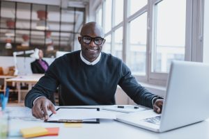 Portrait of confident young man at his desk with laptop doing paperwork. Happy african man looking at camera while at work.